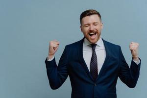 Overjoyed triumphing businessman clenches fists with triumph celebrates successful deal dressed in formal clothes isolated over blue background copy space for your advertisement exclaims from joy photo