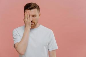 una toma horizontal de un hombre cansado y serio hace que la palma de la cara se sienta aburrida pasando tiempo solo cubre los ojos con la mano vestida con una camiseta blanca informal aislada sobre un espacio en blanco de fondo rosa en el lado derecho foto