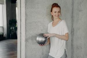 Red haired positive woman standing against gray wall with fitball in hand before pilates class photo