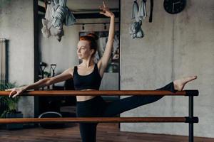 Young athletic red haired ballerina stretching leg on ballet barre in fitness studio photo