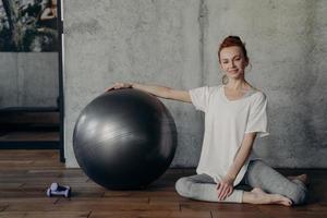 Happy young fitness woman relaxing after pilates workout while sitting with large exercise ball on floor photo