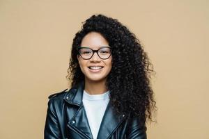 Stylish African American woman smiles toothily, has pleasant talk, enjoys good day, wears transparent glasses, leather jacket, expresses positive emotions, poses indoor at studio. Ethnicity, fashion photo