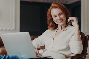 Indoor shot of cheerful satisfied woman works remotely, busy with distance job, looks positively at camera before start working, uses modern laptop computer and wireless internet, browses information