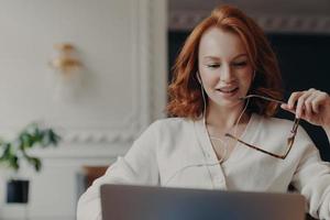 Pleased redhead woman student watches training webinar, has online video conference, uses earphones, works on business project, keeps transparent glasses in hand. Distance work via internet. photo