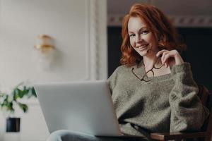 Self confident cheerful redhead woman holds spectacles for vision correction has remote job busy with online project wears warm sweater, sits in front of laptop computer, makes plan of effective work photo