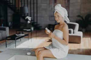 Horizontal shot of attractive lovely woman sits against cozy room background applies body lotion after taking bath uses cosmetic product for glowing skin wrapped in towel undergoes beauty treatments photo