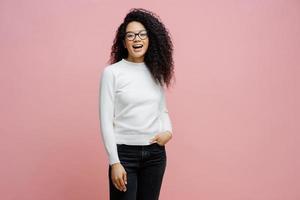 Indoor shot of pretty cheerful woman going to work, laughs at something positive, wears neat white jumper and jeans, keeps hand in pocket, has fun with friends during free time, poses indoor