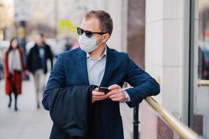 Prosperous businessman in formal wear poses at street, waits for someone, holds mobile phone and sends text messages, wears medical mask during coronavirus outbreak, few people walking outside