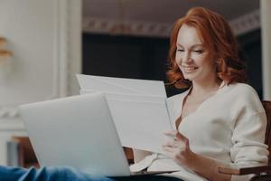 Horizontal view of pleased adult woman entrepreneur works from home, works remotely from home, looks through paper documents, uses laptop computer, wears casual jumper, poses in own cabinet.