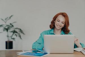 toma horizontal de una mujer independiente de jengibre feliz que trabaja en una computadora portátil en casa, estudia en línea, usa software, navega por información en Internet, se sienta en el escritorio, sonríe positivamente, trabaja de forma remota