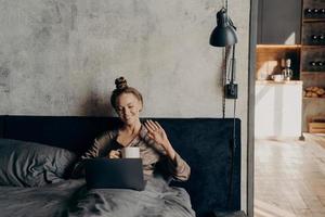 una joven tranquila y relajada acostada en la cama y charlando en línea con sus amigos en una laptop foto