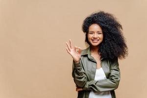 Indoor shot of pleasant looking curly woman has pleasant smile, makes okay gesture, excellent sign, gives approval, dressed in fashionable leather shirt, isolated over brown wall, blank space on left photo