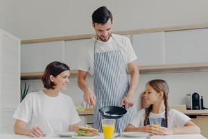 Busy caring father prepares delicious breakfast for wife and daughter, wears apron, puts fried eggs on plate. Cheerful mother and child talk to each other while sit at kitchen table, have meal