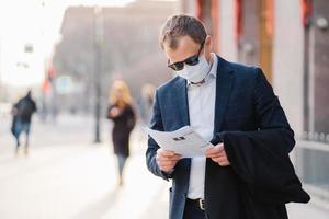 Coronavirus disease. Serious male banker reads newspaper attentively, finds out news about pandemic situation around world, wears protective mask, prevents spreading virus, stands at street.