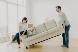 una pareja feliz lleva un sofá blanco moderno con un perro juntos, coloca muebles en la sala de estar, se preocupa por mejorar el diseño interior, comienza a vivir en un nuevo hogar, posa en un apartamento moderno, se divierte foto