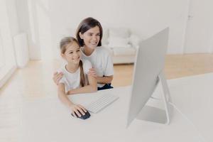 Happy brunette mother helps child do homework on computer, sit together at workplace, have cheerful expressions, focused into monitor. Little girl plays video games via modern gadget with mom