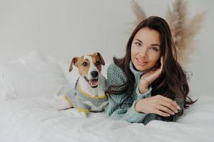 Horizontal shot of pleasant looking tender woman spends free time with favorite pet, looks directly at camera, enjoys domestic atmosphere, cares about dog. People, animals and friendship concept photo