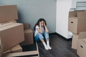 Anxious young lady is sitting on the floor going to relocate. Unhappy hispanic woman packing boxes. photo