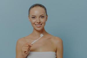 Young happy woman smiling at camera while brushing teeth after shower, enjoying morning routine