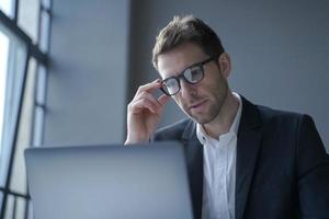 joven empresario alemán concentrado con traje formal y anteojos mirando la pantalla de un portátil