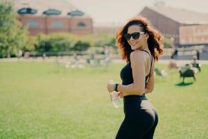 Sport health concept. Sideways shot of happy brunette woman in black top and leggings poses with fresh water wears sunglasses enjoys summer day has regular fitness training. Hydration during workout photo