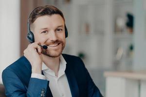 Happy young businessman wearing headphones with mic talking with clients indoors photo