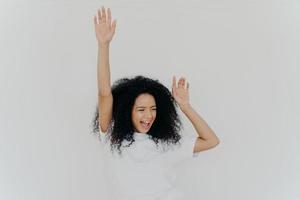 Joyful curly African American woman raises hands up, laughs from happiness, celebrates triumph, rejoices success, wears white t shirt, poses indoor, gestures actively, gets unexpected surprise photo