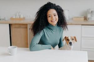 Beautiful curly haired woman dressed in casual turtleneck, sits at white table in kitchen, drinks tea from cup, plays with jack russell terrier dog, enjoys spare time. Afro lady with favourite pet photo