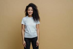 una foto de estudio de una atractiva mujer alegre con peinado afro sonríe positivamente, se regocija comprando ropa nueva, usa pantalones blanco y jeans, disfruta del tiempo libre para descansar, posa contra un fondo marrón