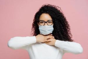 Sick curly haired woman touches neck, suffers from suffocation and shortage of breathing, wears medical mask to avoid virus infection, isolated on pink background. Coronavirus, health care concept photo