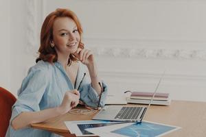 Cute young woman with ginger hair uses laptop computer at home, works from home, sits at desktop, owns online shop, ready to greet clients, gives free consultation, works on research project. photo