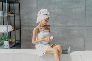 Relaxed young European woman applies moisturizer cream on legs after taking bath sits wrapped in towel in bathroom, enjoys beauty treatments, uses cosmetic product for healthy skin. Hygiene concept photo