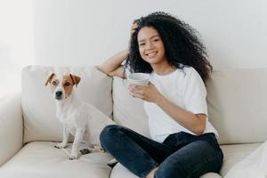 Beautiful woman with Afro haircut, drinks coffee, poses in living room at sofa with pedigree dog, wears white t shirt and jeans, enjoys coziness and comfort at home. People, animals, lifestyle photo