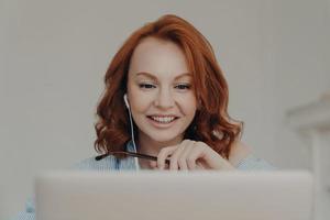 Pretty positive redhead woman gives online consultation, sits in front of opened laptop computer, has discussion with corporate client, smiles happily, uses modern earphones, enjoys online studying photo