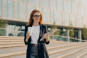 Businesswoman wears elegant formal clothes drinks takeaway coffee carries notebook and modern gadget photo