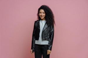 Happy positive Afro American woman dressed in leather jacket, ready for outdoor walk, poses against pink background. Joyful dark skinned millennial girl in good mood has casual talk with friend photo