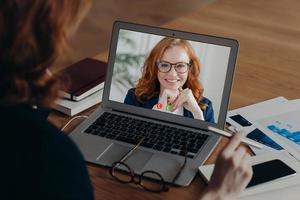 View over shoulder of female freelancer talks with employee by video call, discuss strategy of work. Laptop screen view of smiling ginger woman in spectacles. New technology usage. Distance working