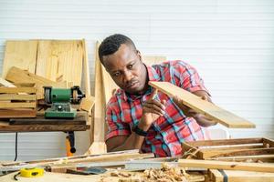 joven carpintero afroamericano mirando y eligiendo madera y usando papel de lija para frotar tablones de madera en la mesa del taller en la fábrica de madera de carpintero