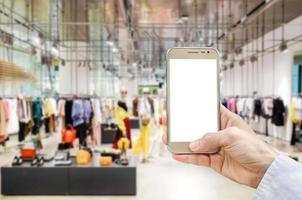 Mockup image of man hand holding gold mobile phone with blank white screen on blurred abstract background of many cotton clothing on the shelves of fashion shop.business concept.shopping online. photo