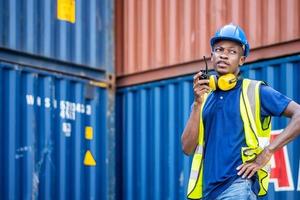 Adult African american men worker Check and control loading freight Containers by use radio at commercial shipping dock feeling curious. Cargo freight ship for import export concept photo