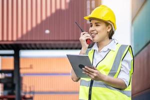 Young asian female engineer or manager holding a Tablet and use of radio communication to control container load in industrial transport and logistics concepts photo