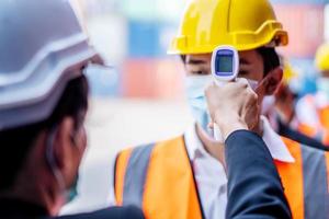 Female foreman wearing a mask measures the temperature for a worker in a safety suit. They stood and waited for the measurement with a non-contact infrared thermometer to prevent covid virus photo