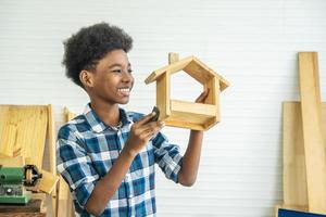 Smiling African-American boy carpenter looks at his own wooden house that helps his father happily do it photo