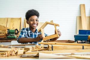 Smiling African-American boy carpenter looks at his own wooden house that helps his father happily do it photo