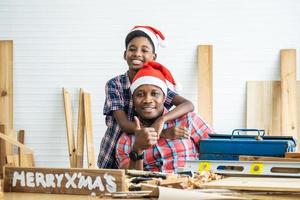 Christmas happy child and father . Cheerful african american son carpenter embracing his father while leaning at the wooden table with diverse working tools laying on it photo