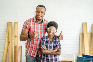 family, carpentry, woodwork, Portrait of cheerful african american father and little son making thumbs up at workshop, with diverse working tools laying on it photo