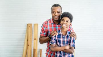 family, carpentry, woodwork, Portrait of cheerful african american father and little son standing by workplace in workshop, with diverse working tools laying on it photo