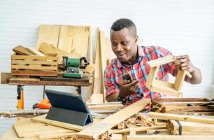joven carpintero afroamericano sentado en la mesa mostrando artículos de madera a través de una tableta mientras chatea en video en línea con el cliente o enseña en línea en la fábrica