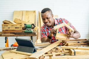 Young african american male carpenter sitting at table showing wooden items through tablet while video online chatting with customer or teaching online in factory photo