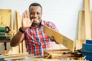 Young african american male carpenter sitting at table showing wooden items through tablet while video online chatting with customer or teaching online in factory photo